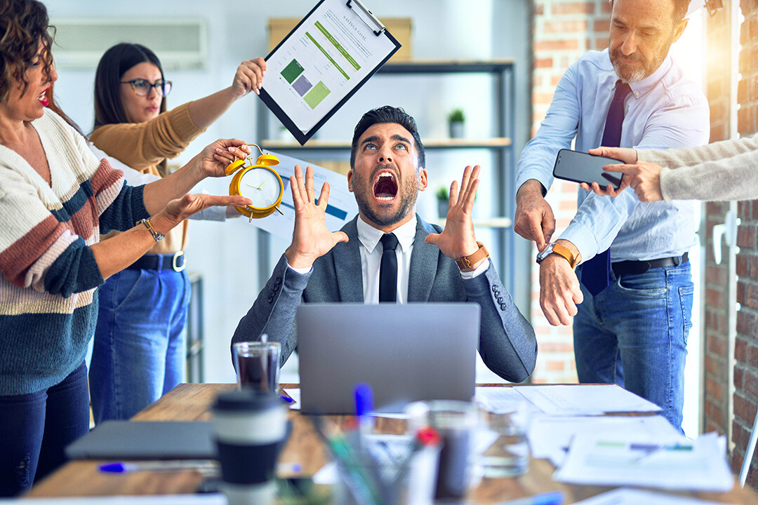 Gestresster Manager im Büro, umgeben von Kollegen, die ihm Druck machen und Aufgaben präsentieren